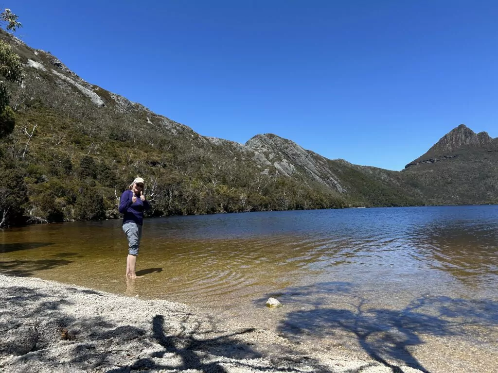 A woman stands in cool waters on the shore of Dove Lake without shoes on.