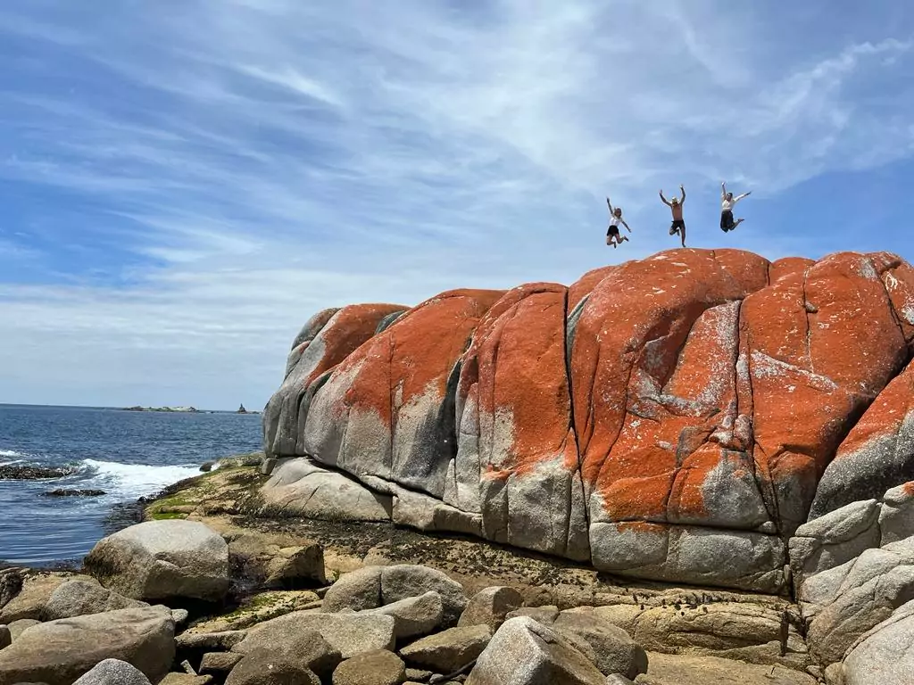 Three people jump for joy on top of large orange bounlders at the Bay of Fires